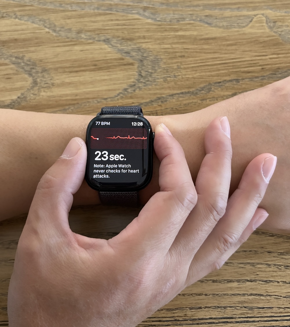 A hand holding an Apple Watch with two fingers during an ECG recording, index finger on the Digital Crown, thumb steadying the watch body
